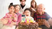 A happy dark-haired child surrounded by family members sits in front of his chocolate birthday cake.