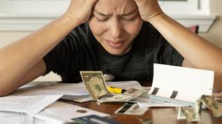 A woman puts her hands on her head in frustration while looking down at a desk scattered with papers and a $1 bill.